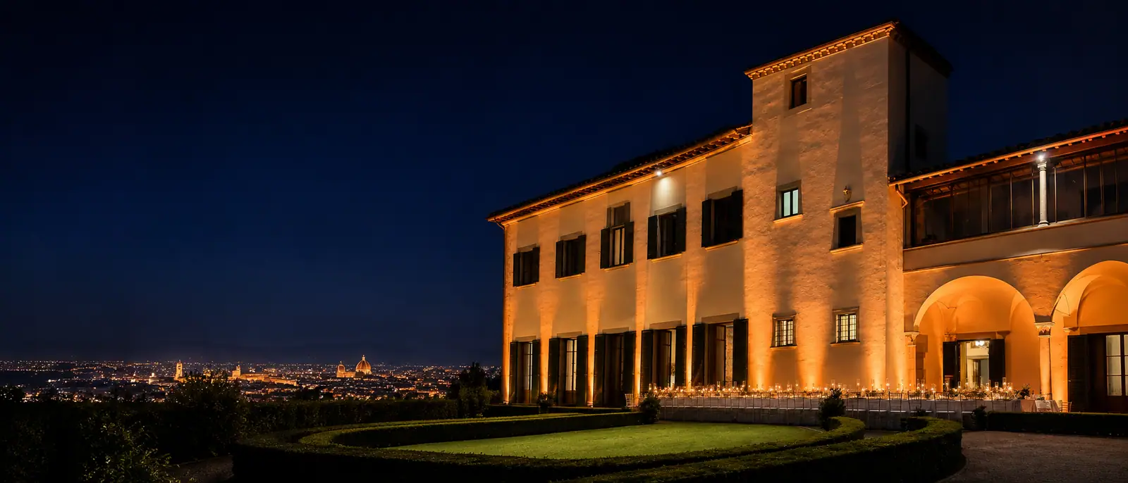 La villa illuminata di notte con la lunga tavolata a lume di candela lungo la facciata, il giardino di bossi in primo piano e Firenze accesa in fondo con la cupola del Duomo
