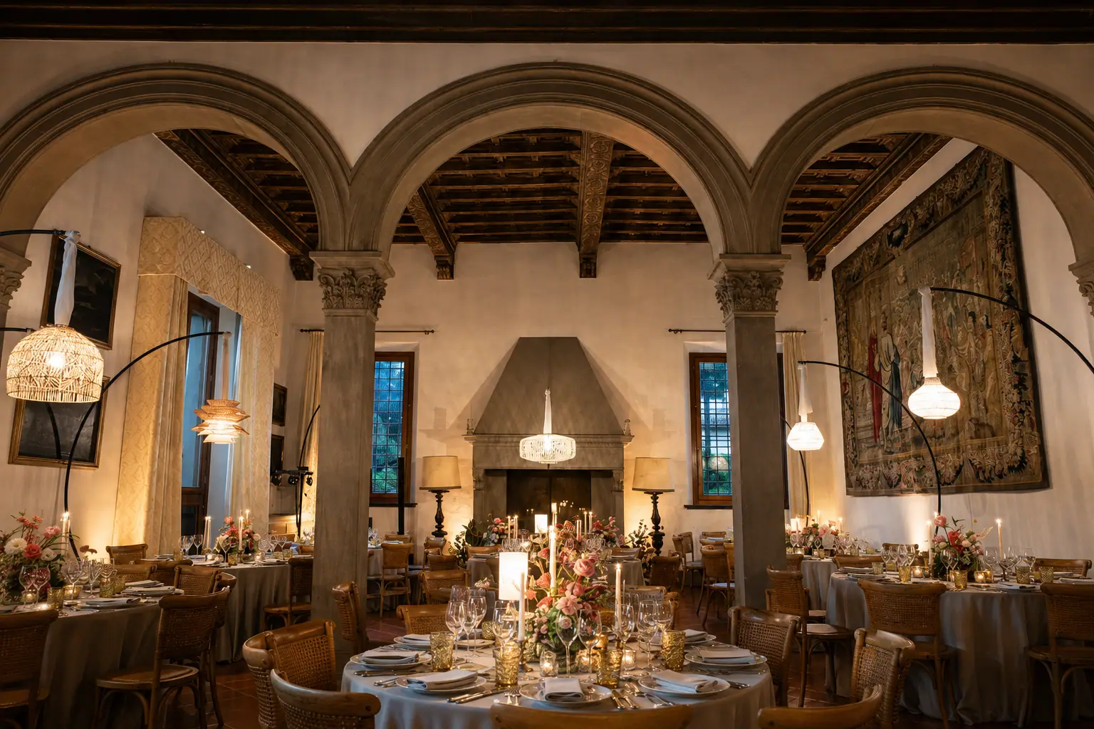 Il salone con il camino apparecchiato per un ricevimento: archi in pietra, soffitto a travi, un arazzo alla parete e i tavoli rotondi con fiori e candele